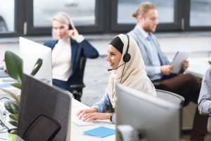 selective focus of smiling arabic call center operator in hijab in headset working on computer at